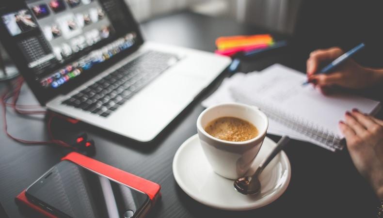 A desk arrangement at home featuring a cup of coffee, a laptop and a woman taking notes.